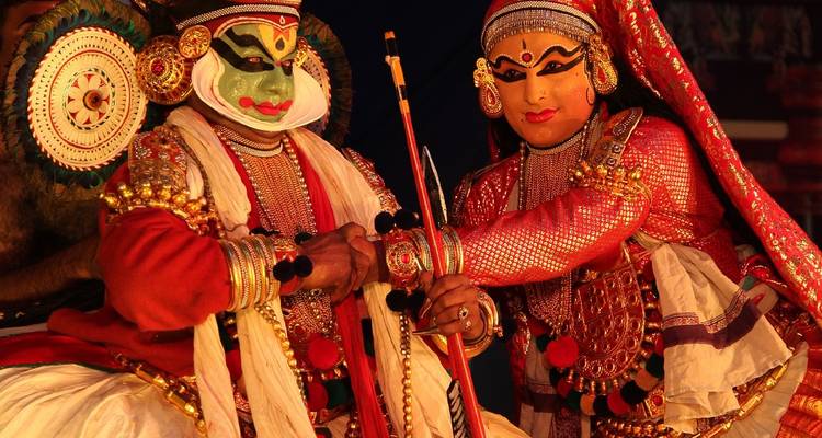 Two elaborately costumed Kathakali performers in vibrant red and gold attire pose on stage with dramatic makeup and props.
