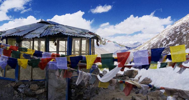 Colorful prayer flags flutter at a snowy high-altitude mountain pass shelter.