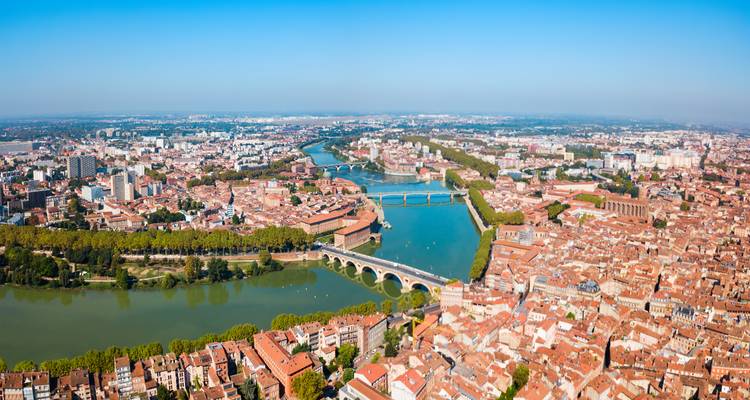 Weitläufige Luftaufnahme von Toulouse mit der Garonne, mehreren Brücken und terrakottafarbenen Dächern, die sich bis zum Horizont erstrecken.