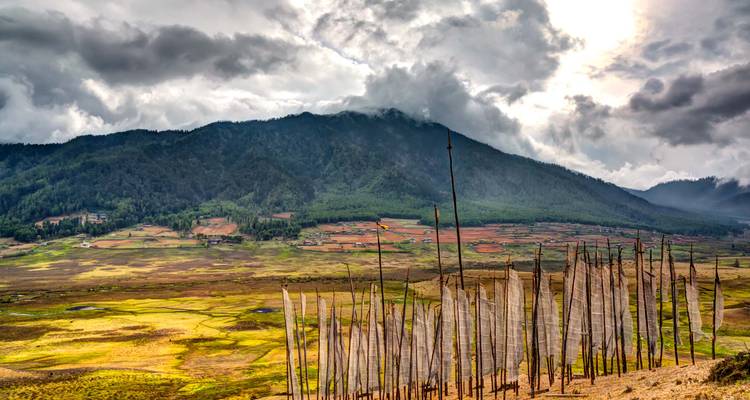 Breites Tal, übersät mit Gebetsfahnen und einem Flickwerk aus Feldern unter düsteren Wolken und bewaldeten Gipfeln