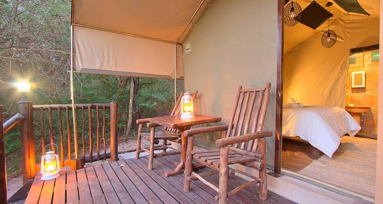 Leinwand-Safari-Zelt mit hölzernen Schaukelstühlen und Laternen auf einer erhöhten Terrasse in der Abenddämmerung.