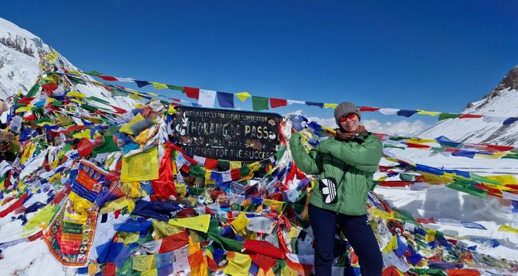 Smiling trekker poses beside prayer-flag-covered Thorong La Pass sign in snowy Himalayas.