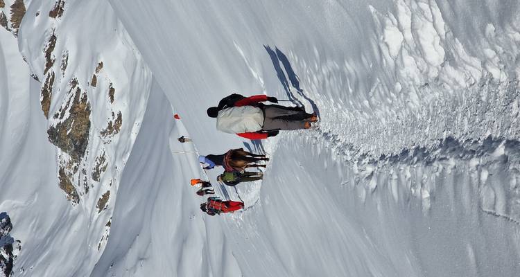 Line of climbers and pack horses ascend a narrow snowy trail between white slopes.