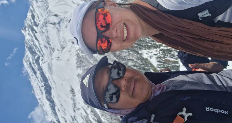 Close-up selfie of two trekkers in sunglasses with snowy peak behind them.