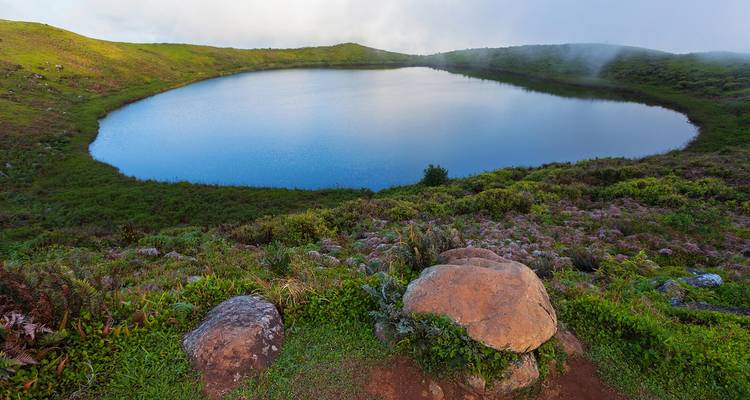 Lac de cratère parfaitement circulaire entouré de collines vertes brumeuses dans la douce lumière matinale.