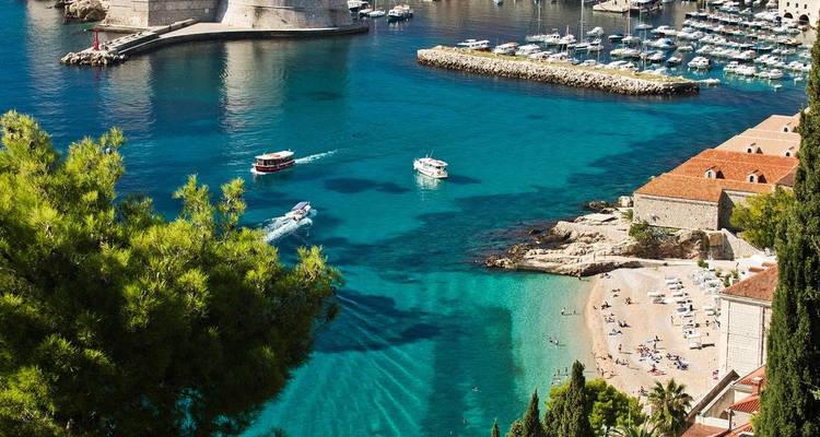 Turquoise Adriatische baai onder de stadsmuren van Dubrovnik met kleine bootjes en zonnebaders op een zandige inham.