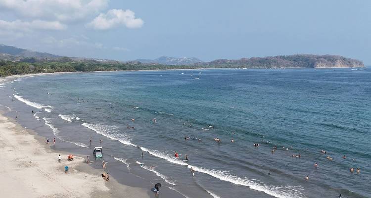 Wide sandy beach and blue ocean bay with dozens of swimmers and sunbathers scattered along the shoreline.