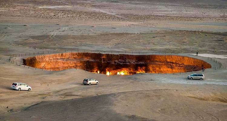 Wide desert plain with the fiery Darvaza gas crater glowing orange at dusk, a few vehicles parked nearby.