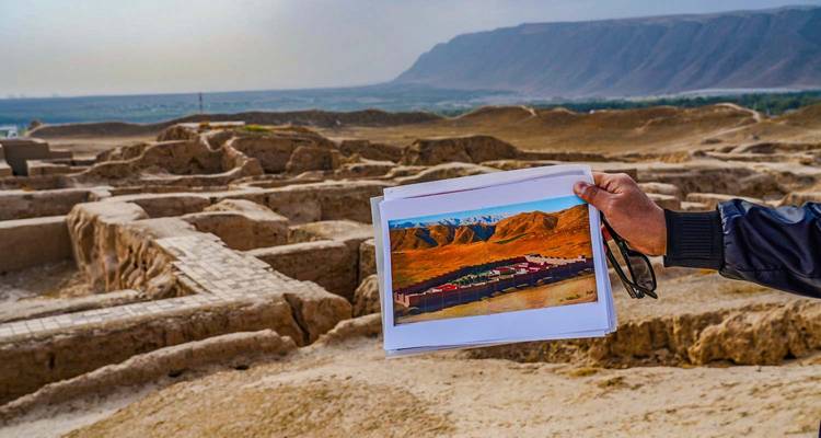 Hand holding a historical reference photo over ancient adobe ruins with distant mountains in hazy light.