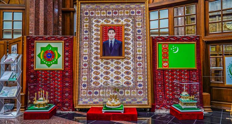 Indoor ceremonial display featuring a framed presidential portrait flanked by Turkmen flags and ornate carpets.