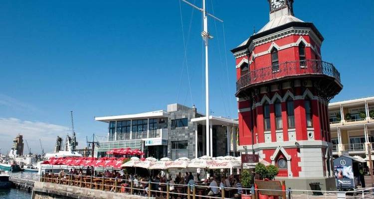 Red Victorian clock tower and waterfront restaurants bustling with diners under a bright sky.