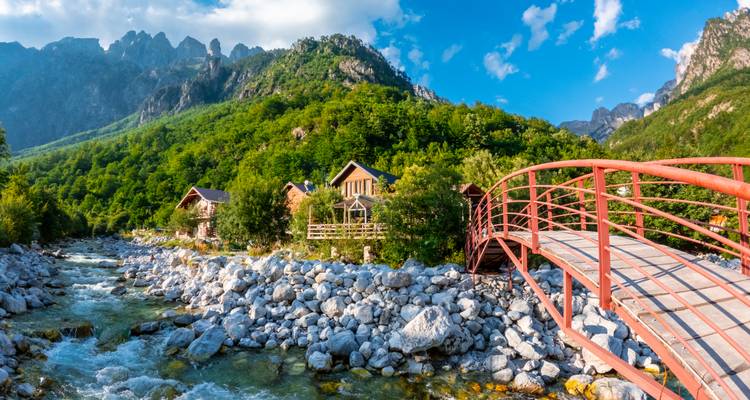 Red pedestrian bridge over a rocky mountain stream leading to wooden chalets nestled in lush green forested hills.
