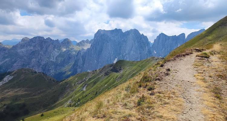 Mountain hiking trail climbing along a grassy ridge with towering jagged peaks under partly cloudy skies.