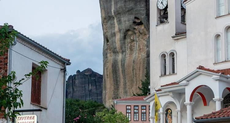 Estrecha calle del pueblo con el imponente pilar rocoso de Meteora elevándose detrás de edificios encalados.