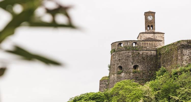 Torre de reloj de piedra sobre una muralla fortificada que se alza por encima del follaje verde de la ladera contra un cielo nublado.