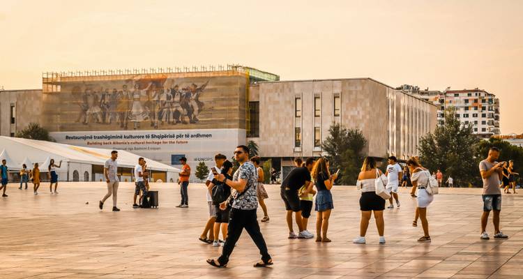 Locals and tourists mingling on the expansive paved square before Tirana’s National History Museum at sunset glow.