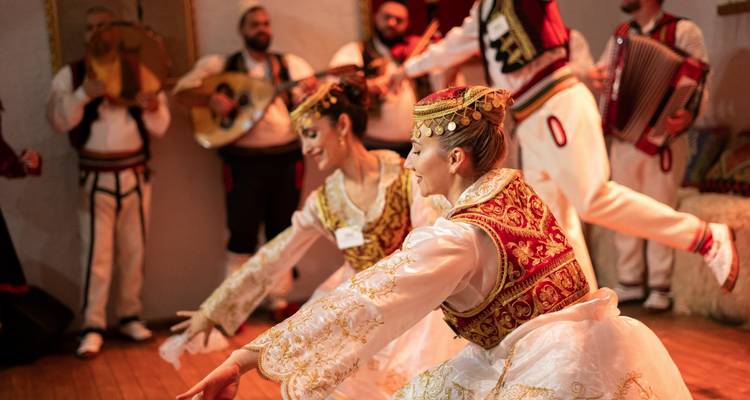 Traditional dancers in ornate Albanian costumes perform energetically indoors.