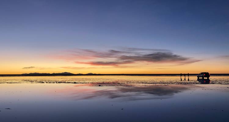 Atardecer sereno en el Salar de Uyuni con colores pastel del cielo reflejados perfectamente en el agua poco profunda, un vehículo solitario y siluetas en la distancia.