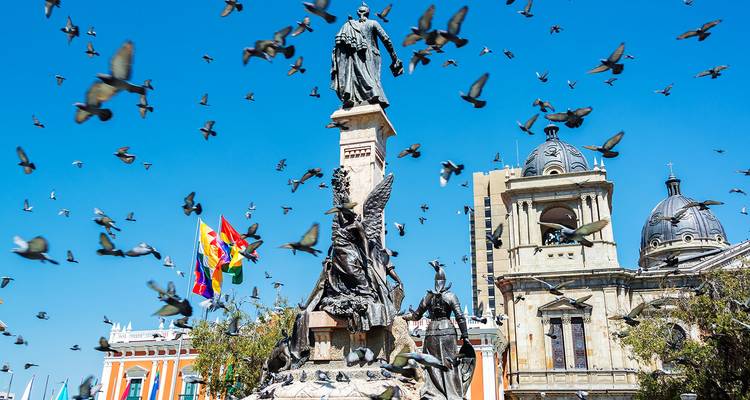 Zentralplatz von La Paz mit Tauben, die um Statuen und bolivianische Flaggen gegen einen strahlend blauen Himmel fliegen
