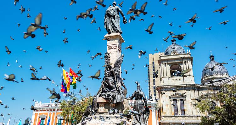 A historic plaza with towering monument, Bolivian flags, and swirling pigeons under a bright blue sky.