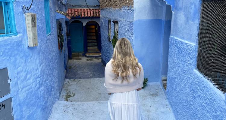 A woman in a white dress walks down a quiet blue-washed alley toward an arched doorway.