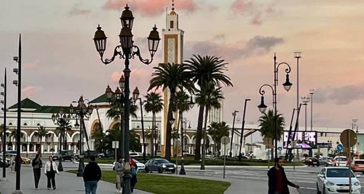 People cross a modern plaza lined with palms and ornate street lamps in front of a white mosque at sunset.
