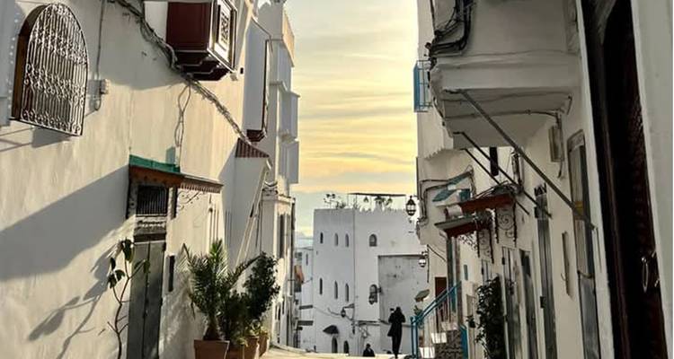 Narrow whitewashed lane with wrought-iron balconies and evening light filtering in while a couple walks below.