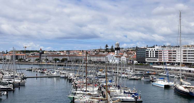 Drukke jachthaven vol zeilboten aan de compacte waterkant skyline van Ponta Delgada onder een gedeeltelijk bewolkte hemel.