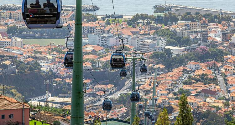 Line of cable cars ascending above Funchal city with Atlantic coastline and red rooftops below.