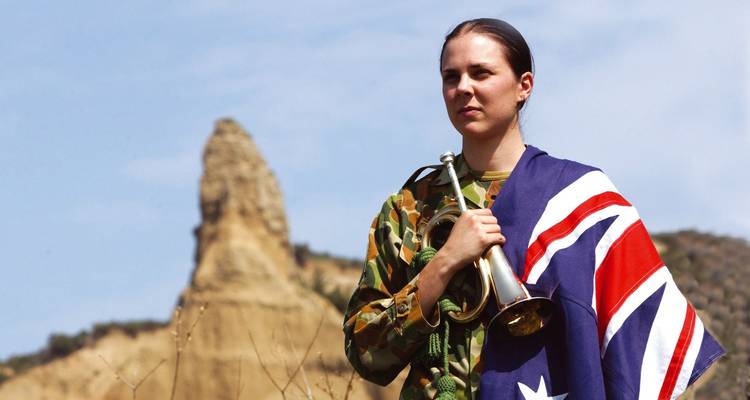 Young bugler draped in Australian flag standing at Gallipoli battlefield ridge.