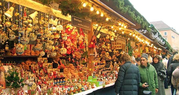 Bustling row of Christmas-market stalls overflowing with ornaments and treats while shoppers browse beneath glowing bulbs.