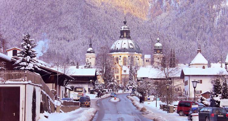 Snow-covered alpine village road leading toward a domed baroque church framed by forested mountains glowing in winter light.
