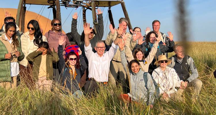 Grupo de turistas emocionado posa con las manos levantadas junto a la canasta de un globo aerostático aterrizado en un campo abierto.