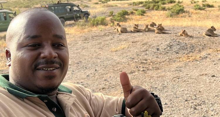 Guía se toma una selfie haciendo el gesto de pulgar arriba con leones descansando en un camino de tierra detrás.
