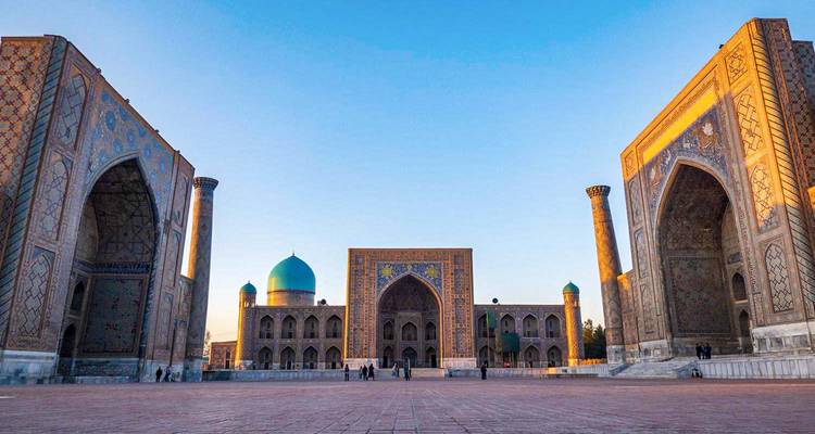 Vista de ángulo amplio del amanecer en la Plaza Registan bañada por la cálida luz del alba, con solo algunos visitantes en la inmensa plaza.