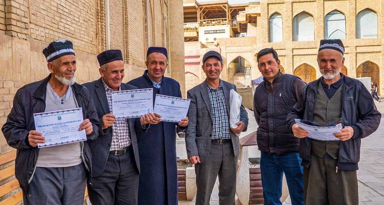 Grupo de hombres mayores sosteniendo orgullosamente certificados afuera de una pared histórica de ladrillo de una madrasa.