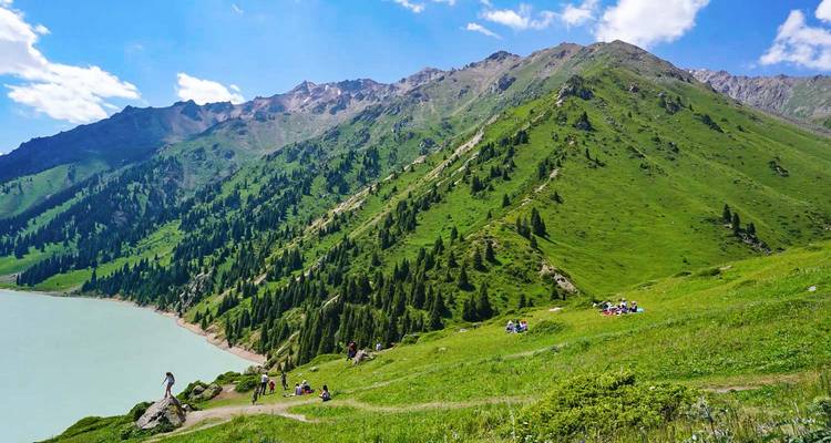 Ladera alpina cubierta de hierba que desciende hacia un lago azul pálido con pícnickers y excursionistas dispersos bajo picos escarpados.