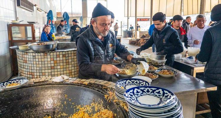 Bullicioso comedor donde un chef sirve plov de un enorme kazan rodeado de pilas de platos con patrones azules.
