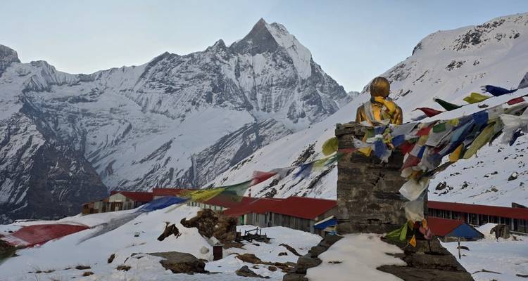 A golden statue wrapped in prayer flags overlooks red-roofed lodges beneath a jagged Himalayan peak.