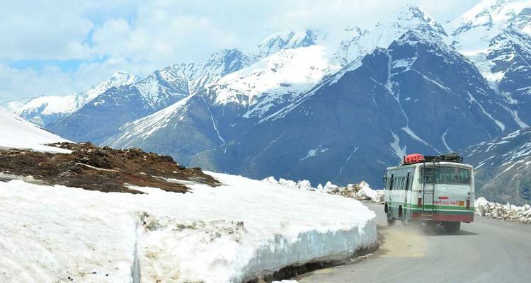 A Himachal road bus winds along a snow-walled mountain pass flanked by towering, snow-capped peaks.