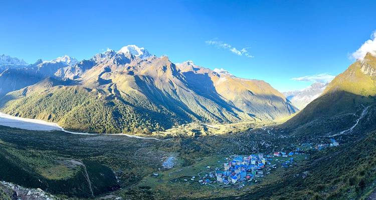 Vue panoramique d'une large vallée alpine avec un groupe de chalets aux toits bleus sous d'imposants sommets enneigés.
