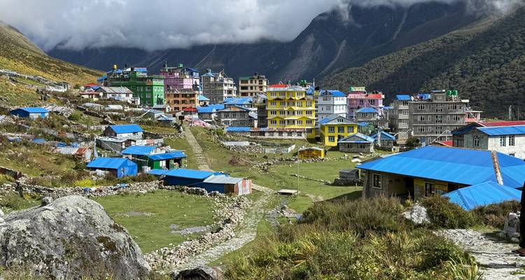 Des bâtiments colorés à plusieurs étages parsèment un village himalayen niché entre des collines verdoyantes et des nuages bas.