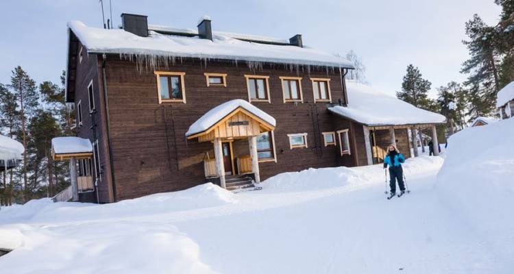Skiër glijdt voorbij een houten lodge in diepe sneeuw onder winterzonlicht.