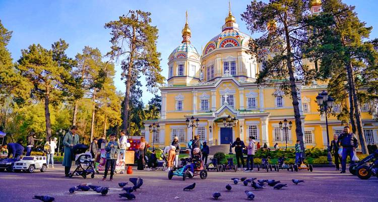 Les familles et les enfants nourrissent les pigeons devant la somptueuse cathédrale Zenkov de couleur jaune, entourée de grands sapins.
