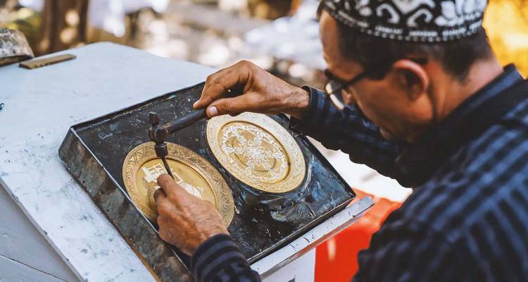 Un artisan grave soigneusement des motifs ornés sur une plaque de métal doré à l'aide d'un petit marteau et d'un ciseau.
