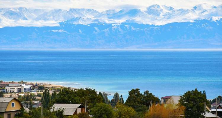 Le lac Issyk-Kul d'un bleu profond s'étend vers des montagnes aux pics enneigés avec un village en bord de lac au premier plan.