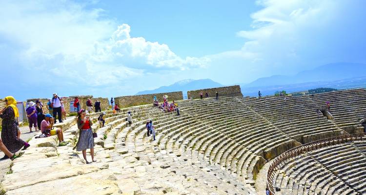Bezoekers verkennen een uitgestrekt stenen amfitheater met uitgestrekte rijen zonbeschenen antieke zitplaatsen en uitzicht op de bergen.