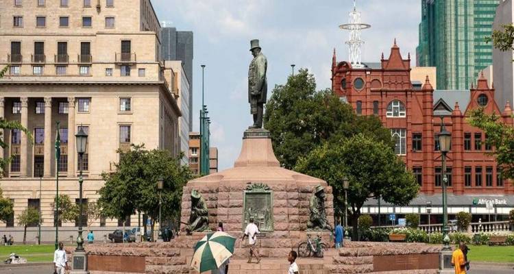 Central Joburg square with historic statue on stone plinth, flanked by classic and modern buildings.