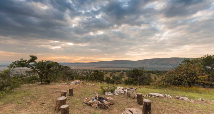 Vaste panorama de savane avec arbres épars, foyer de pierre et ciel dramatique de lever de soleil nuageux.