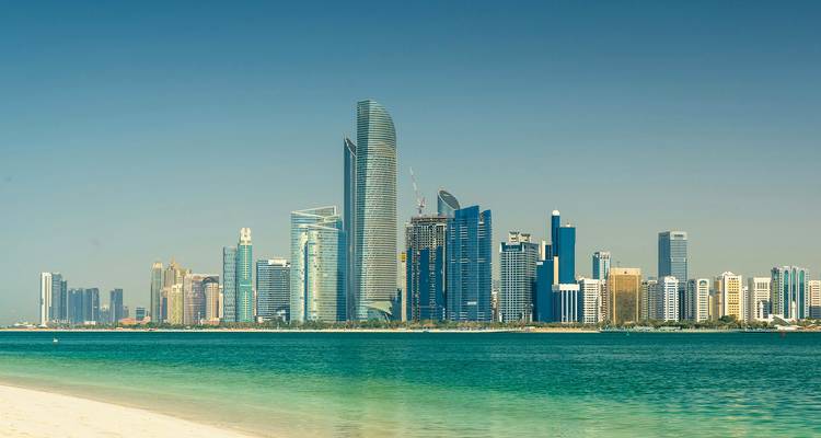 Modern skyline of glass skyscrapers rising behind turquoise Gulf waters and white sand beach.